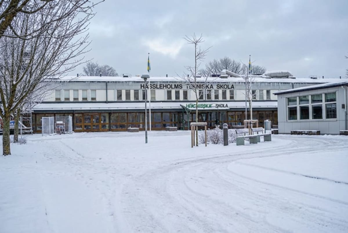 En fasadvy av en skolbyggnad, Hässleholms Tekniska Skola, under vintertid. Byggnaden är en lång, enplansbyggnad med stora panoramafönster i markplan och mindre, rektangulära fönster på övervåningen. Taken på byggnaderna är snötäckta och isstycken hänger från takkanten. Framför skolan finns ett snötäckt landskap med smala gångvägar eller vägar som slingrar sig genom snön. Till vänster i bild syns grenar från ett barrträd täckta med snö. Mot mitten framför byggnaden står flera smala, kala träd och det finns några bänkar och lyktstolpar i förgrunden och närmare byggnaden. Två flaggstänger med svenska flaggor sträcker sig upp mot en grå, molnig himmel. Till höger om huvudbyggnaden finns en mindre byggnad med stora fönster. Marken är helt täckt av snö.