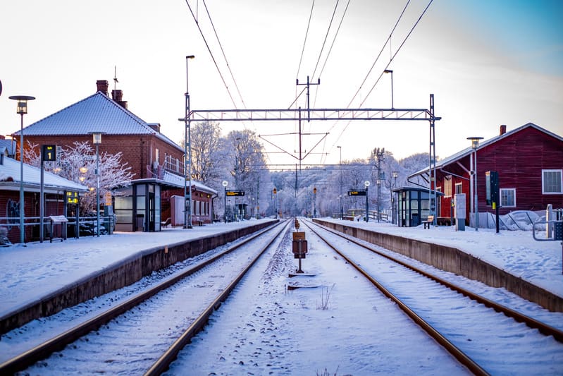 Bilden visar en järnvägsstation täckt av snö under vad som ser ut att vara en kylig vinterdag. Två spår sträcker sig ut från förgrunden i bildens mitt mot horisonten. På vardera sida om spåren finns snötäckta perronger. Till vänster syns ett större t