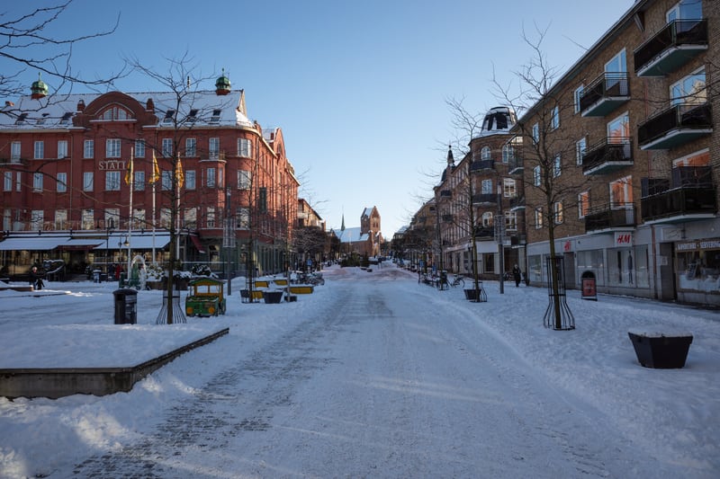 En gata täckt av snö. På den vänstra sidan av gatan finns en stor röd byggnad med texten "STATT" på fasaden, flaggor hänger utanför och den har flera fönster och balkonger. På höger sida av gatan finns en ljusare tegelbyggnad med balkonger och butiks