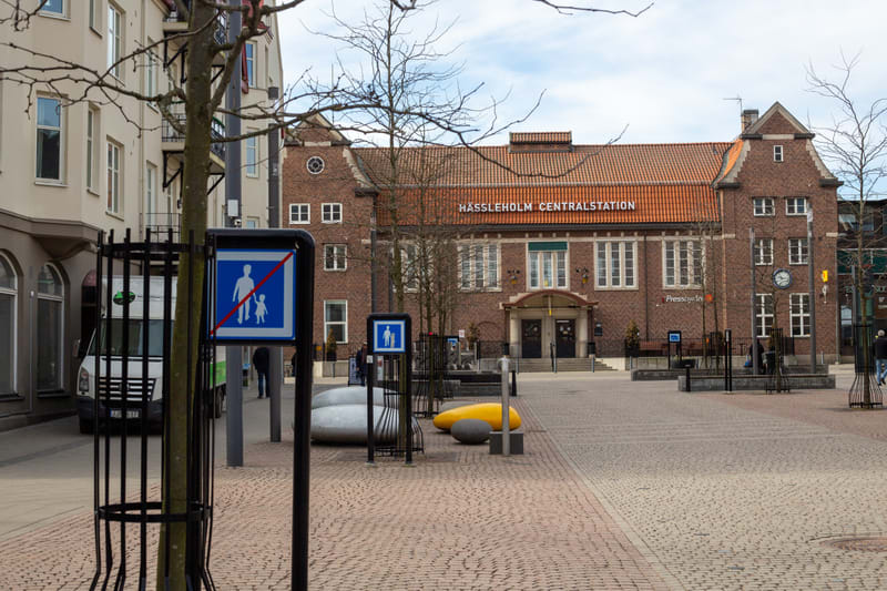 Bilden visar ett torg framför Hässleholms Centralstation. Centralstationsbyggnaden är uppförd i tegel med ett rött tegeltak och har texten "HÄSSLEHOLM CENTRALSTATION" skrivet i orange färg på fasaden. Till vänster om stationen syns delar av en ljus b