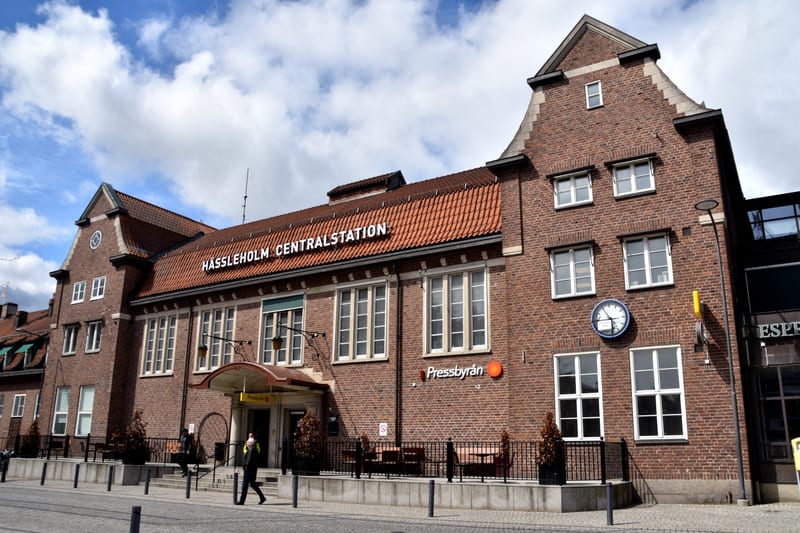 En bild som visar framsidan av en tegelsbyggnad med ett textskylt som lyder "HÄSSLEHOLM CENTRALSTATION" på taket. Byggnaden har många fönster med vita karmar och ett rött tegeltak. Till vänster om ingången finns en skylt med texten "Pressbyrån". Det