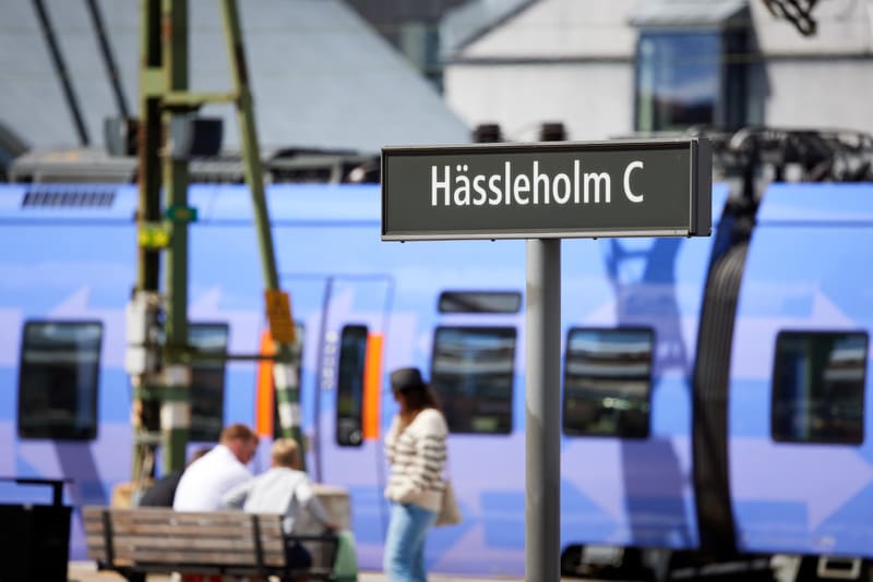 Bilden visar del av en järnvägsstation med ett skylt i förgrunden där det står "Hässleholm C" med vit text på en mörk bakgrund. Skylten är monterad på en grå stolpe. Bakom skylten och suddigt i bakgrunden syns ett blått tåg med rektangulära fönster.