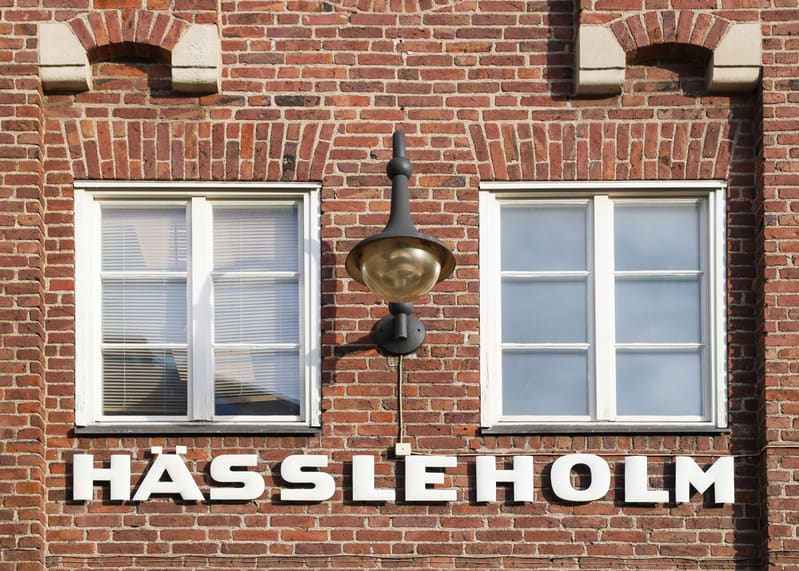 Bilden visar delar av en tegelbyggnads fasad. Centralt i bilden är det vita texturet HÄSSLEHOLM monterat på tegelväggen. Ovanför texten finns två vitmålade fönster med spröjs. Det vänstra fönstret har genomskinliga persienner synliga genom glaset, me