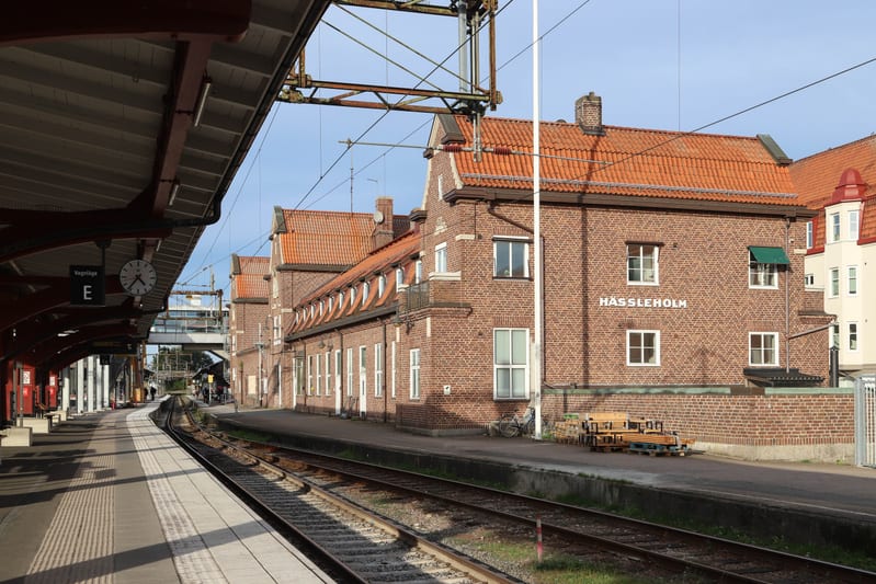 Bilden visar en järnvägsstation från en av perrongerna. Till vänster i förgrunden syns taket på perrongen, med en klocka och en skylt där det står "Vägval E". Spåren löper i bildens centrum, och till höger om spåren ligger en byggnad med fasad av röt
