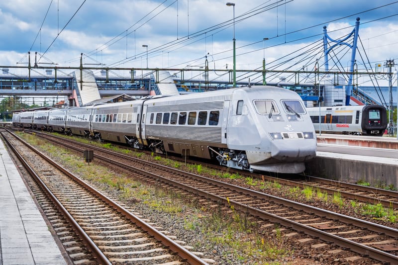 Bilden visar ett tåg uppställt vid en tågstation under dagtid med delvis molnig himmel. I förgrunden till vänster syns delen av en perrong. Flera parallella järnvägsspår sträcker sig genom centrala bilden, med gräs och grus mellan spåren. Till höger