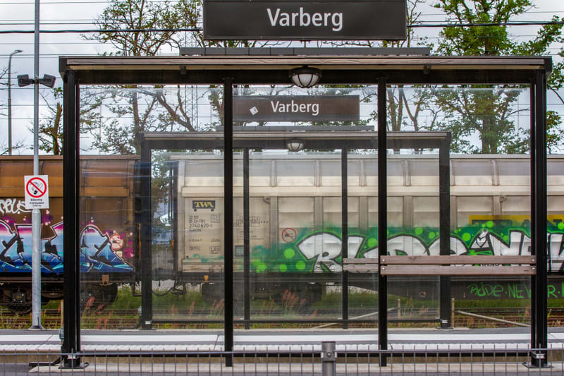 En bild som visar en tågperrong med en väntkur i förgrunden. Ovanför väntkuren hänger en stor svart skylt med vit text som visar ortsnamnet "Varberg". In genom den transparenta väntkuren syns ett stillastående godståg med flera vagnar. Tågvagnarna är