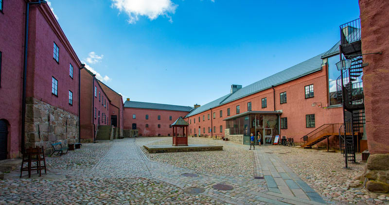 Bilden visar en innergård med kullerstensbeläggning, omgiven av flera byggnader i varierande rödaktiga nyanser. På den vänstra sidan av innergården står ett par trästolar och en bänk, placerade intill en byggnads bas som består av stora stenblock. By