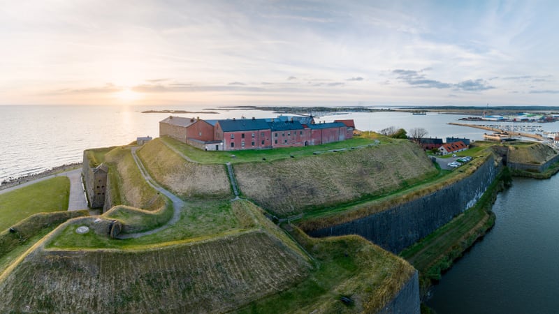 En vidvinkelbild tagen från ovan, som visar en stor, röd tegelbyggnad belägen på en gräsbevuxen kulle omgiven av vatten. Byggnaden är en del av en befästning med vallar och kanaler som omger den. Till vänster i bilden syns ett hav med en horisont där