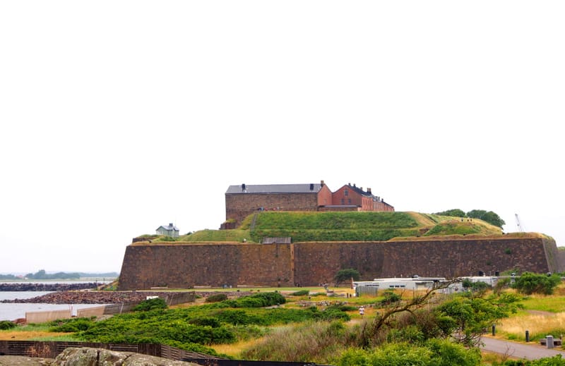Bilden visar ett stort befästningsverk eller en borga som är omgiven av gräsklädda jordvallar. Byggnaden på toppen av höjden är till större delen byggd i mörk sten med ett mörkt tak. Till höger om stenbyggnaden syns ett antal mindre röda byggnader me