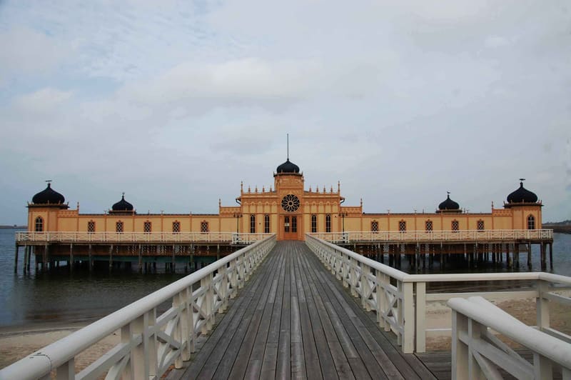 Bilden visar Varbergs Kallbadhus, en gul byggnad med orientaliska inslag, belägen över vatten. En träbrygga leder fram till byggnaden, där mittenpartiet har en stor rund fönsterdekoration och kupoltak med en spira. Byggnaden har flera svarta kupoler.