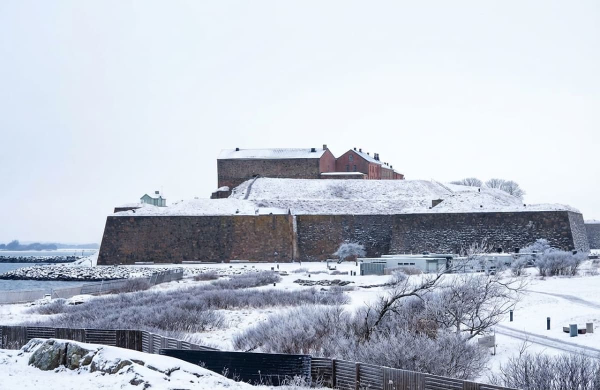 En landskapsbild tagen under dagtid som visar en snötäckt befästning med stora stenmurar. Ovanpå befästningen syns flera rödtonade byggnader med mörka tak, delvis täckta av snö. En liten grön byggnad är också synlig på den övre delen av befästningens mur. Framför befästningen finns ett snötäckt landskap med buskage och kala träd som är täckta av frost. Nederst i bilden syns en del av ett staket, och lite till vänster djupsyn ett vattenområde med en stenig strandlinje. Hela himlen är ljus och molnig, vilket bidrar till ett diffust ljus.