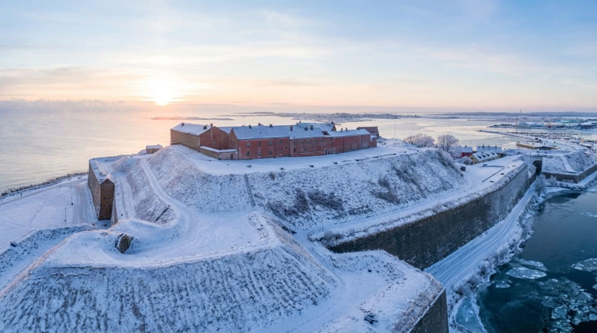 En vidvinkelbild tagen från ovan visar ett historiskt befästningsverk täckt av snö. En stor del av bilden upptas av de snötäckta vallarna och byggnader i rött tegel som reser sig över landskapet. Sjön i bakgrunden är delvis istäckt med isflak som flyter på vattnet. Solen står lågt vid horisonten och skapar ett mjukt, diffust ljus över vattnet och himlen. Till höger i bilden ses en del av en hamn eller en industriområde. Hela scenen är täckt av ett vitt snötäcke, vilket indikerar att bilden är tagen under vintertid. Den ljusa solen i bakgrunden skapar en silhuett av förgrunden.