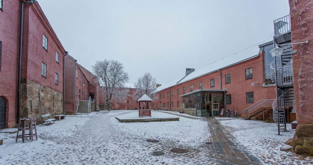 En vidvinkelbild tagen på en borggård täckt av snö. Byggnaderna som omger borggården är tegelröda och har flera fönster. Den vänstra byggnaden har en stengrund. På borggården syns stenläggning som delvis är snötäckt, och gångar i rätblocksstensättning leder genom snön. Till vänster i bilden står vad som ser ut att vara två trästolar och en bänk, delvis täckta av snö. I mitten av borggården, framför träd med frostiga grenar, står ett litet rött lusthus av trä. Till höger syns en byggnad med en inglasad del i markplan och en utvändig spiraltrappa i metall som leder upp till högre våningar. En snötäckt lampa hänger nära spiraltrappan. Himlen är grå och mulen. Tak och mark är täckta av ett tunt lager snö och frost.