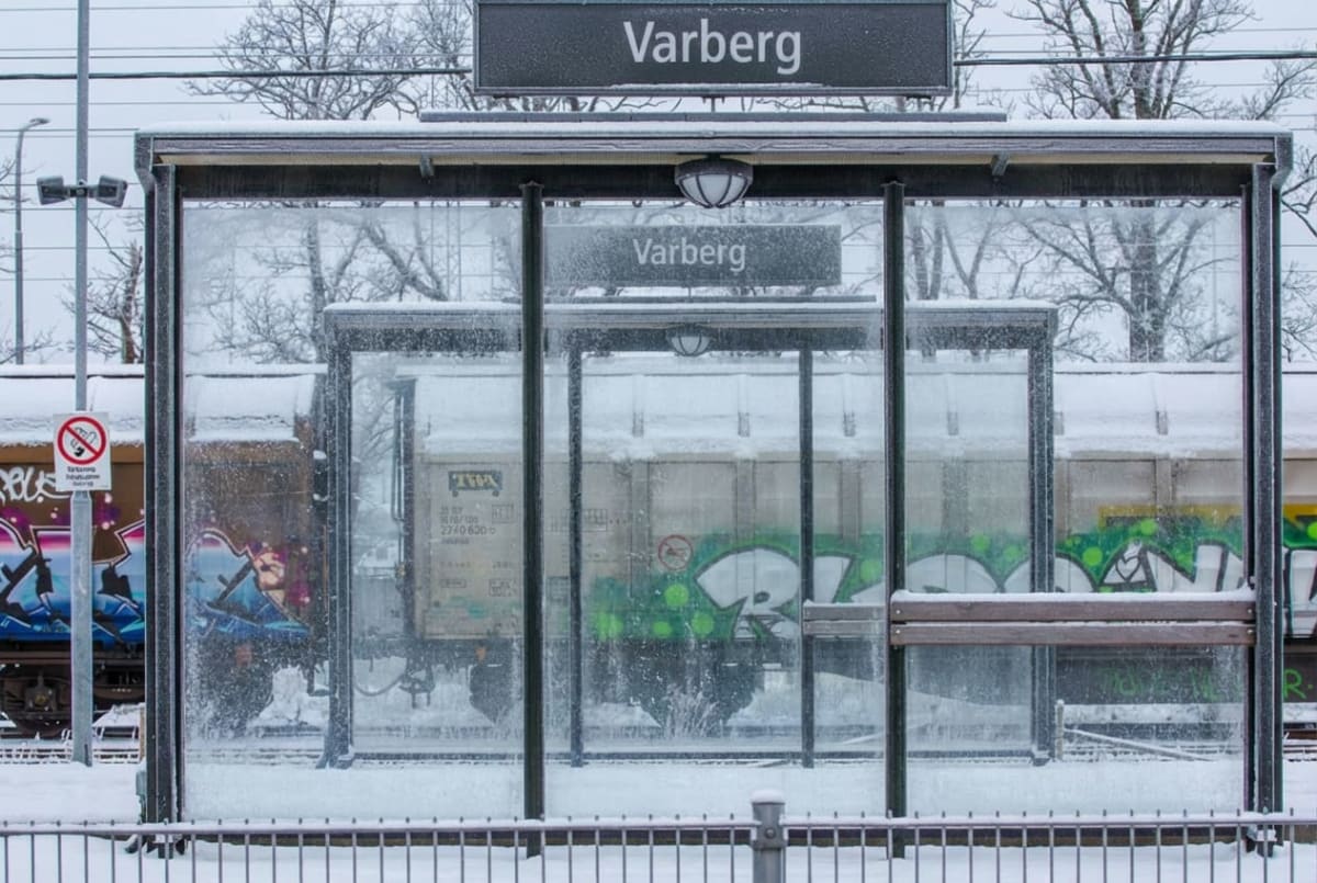 Bilden visar ett snötäckt landskap med ett buss- eller tågskydd i förgrunden, omgivet av snö. Skyddet är huvudsakligen tillverkat av glas och metall, med ett träbänkselement synligt inuti. Över skyddet och även inuti hänger skyltar med texten 'Varberg'. Bakom skyddet syns flera tågvagnar, varav en är täckt med graffiti och en annan är en lastvagn delvis täckt av snö. En varningsskylt med ett överkorsat märke, som ser ut som en person som slänger något, är fäst vid en stolpe till vänster i bilden. Träd med kala grenar och elledningar syns i bakgrunden, allt belagt med snö eller frost. Marken är vit av snö.