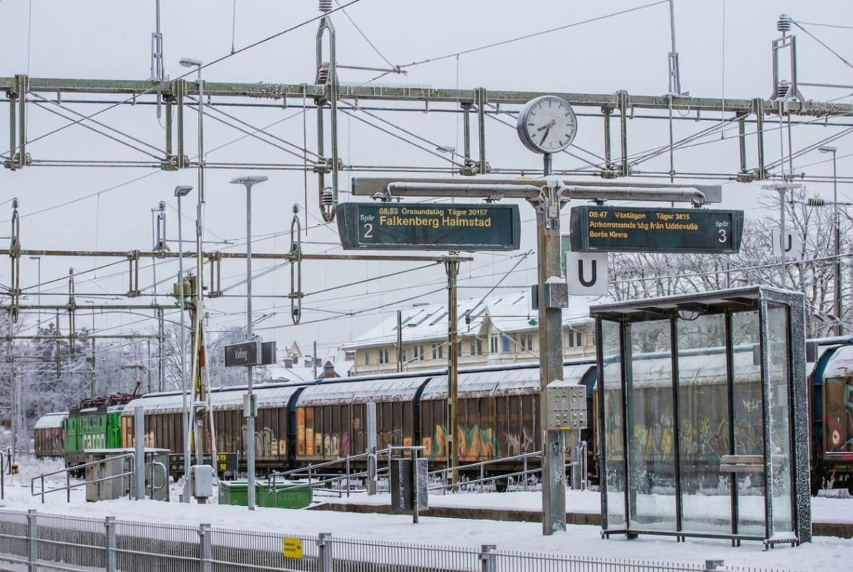 Bilden visar ett snötäckt järnvägsområde med spår, perronger och en tågvagn. Mark och tak är täckta av snö, och frost syns på ledningar och ett busskur-liknande väderskydd. Ovanför spåren hänger ett omfattande nätverk av kontaktledningar och stolpar, delvis täckta av snö och frost. Flera skyltar är synliga, inklusive tåginformationstavlor som visar avgångar till Falkenberg och Halmstad från spår 2, samt information om Västtåget och ankommande tåg från Uddevalla till Borås/Kinna från spår 3. Ett godståg med flera vagnar står på ett av spåren, och delar av vagnarna ser ut att vara täckta av graffiti. En klocka är monterad på en stolpe intill tåginformationstavlorna.