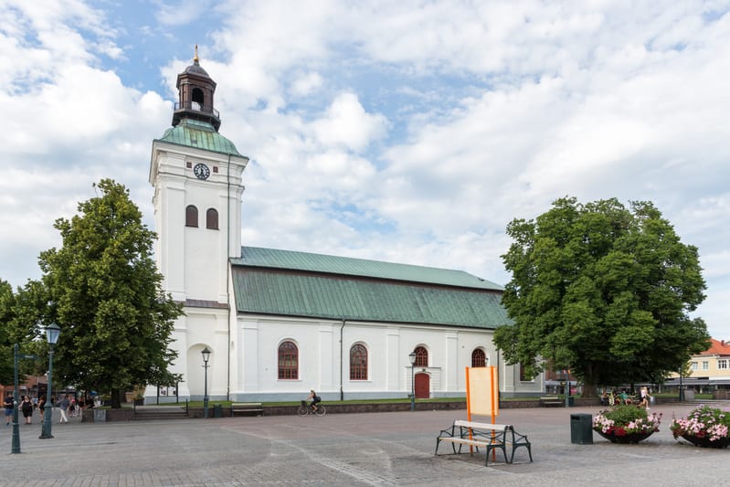 Denna bild visar en kyrka med vit puts och ett klocktorn med ett koppargrönt tak under en molnig himmel. Kyrkobyggnaden har flera runda fönster med röda karmar. Framför kyrkan ligger ett torg belagt med gatsten, där det står bänkar, soptunnor och inf