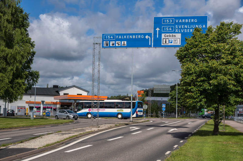 Bilden visar ett vägkors med en vägskylt som pekar mot Falkenberg till vänster och Varberg, Svenljunga, Älvsåker samt Gekås till höger. På vänster sida av korsningen syns en blå- och vitfärgad buss märkt "Shoppabussen" vid en bensinmack med en orange