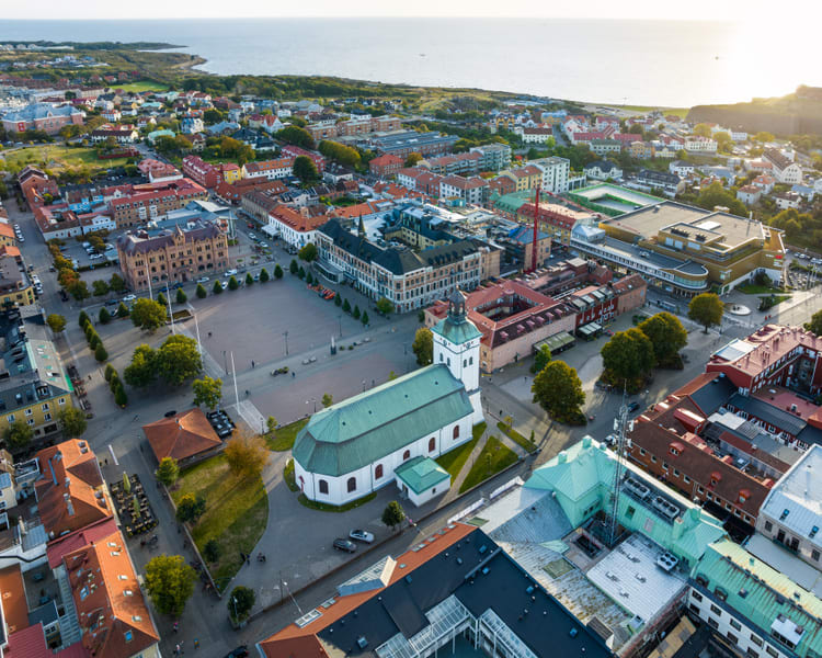 Bilden visar ett flygfoto över en stadskärna med en stor kyrka i förgrunden och havet i bakgrunden. Kyrkan har en vit fasad och ett grönt tak med en klockstapel. Runt kyrkan finns en stor öppen plats, troligtvis ett torg, med träd längs kanterna och