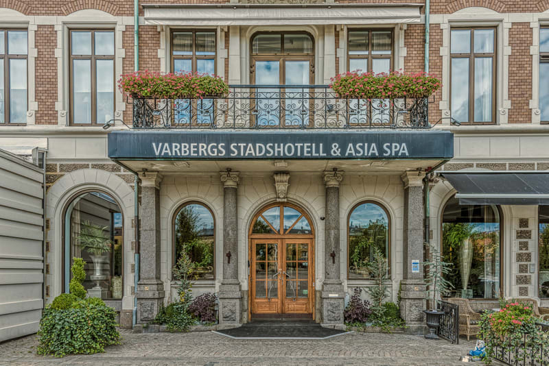 En fasadbild som visar entrén till "VARBERGS STADSHOTELL & ASIA SPA". Byggnaden har en tegelfasad på den övre delen med flera fönster i bruna ramar. Nedanför tegelfasaden finns ett balkongräcke i smide med blommande rosa plantor i blomlådor. Entrén b