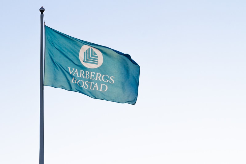 En bild som visar en turkos flagga, Varbergs Bostad. Flaggan är monterad på en flaggstång, vars övre del är synlig i bildens vänstra kant. Logotypen på flaggan består av ett vitt cirkulärt märke med ett stiliserat L-format mönster inuti, samt texten
