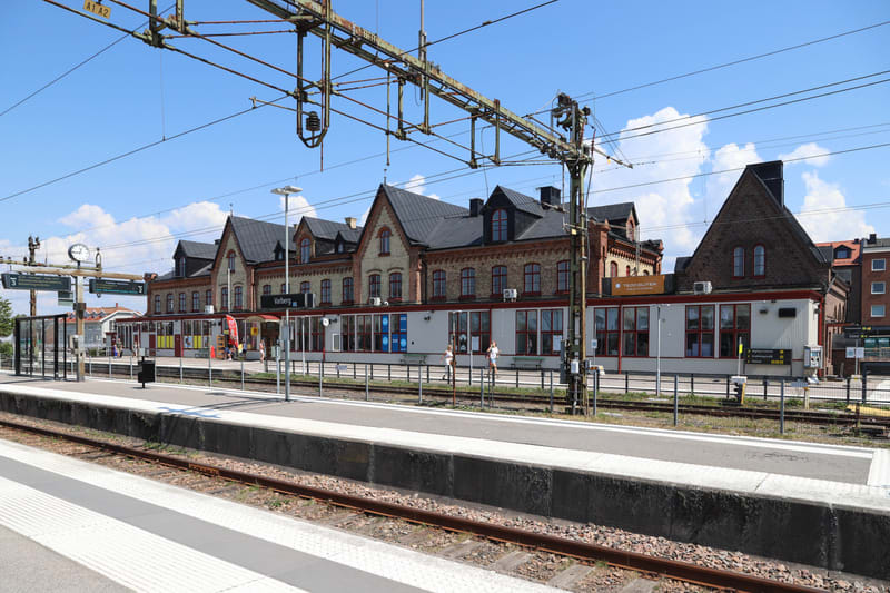 Bilden visar en järnvägsstation från perrongen sett mot stationsbyggnaden. I förgrunden syns räls med vita markeringar på en perrong. Ovanför rälsen och perrongen sträcker sig kontaktledningar för tåg över bilden. Stationsbyggnaden i bakgrunden är te