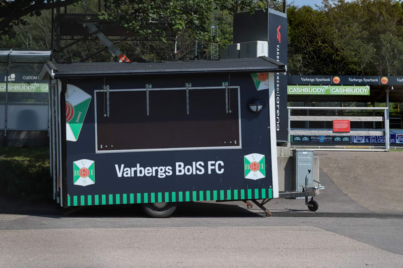 Bilden visar en mörkblå kiosk på hjul med texten "Varbergs BoIS FC" och klubbens logotyp. Kiosken har ett fönster som är täckt och en lampa på sidan. Bakom kiosken syns en del av en läktare med reklam för "Varbergs Sparbank" och "CASHBUDDY". Till vän