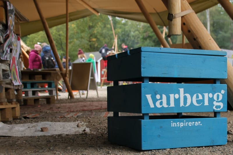 Bilden visar ett utomhusområde under ett tält eller en markis, där fokus är på en turkoslackerad träback i förgrunden. På träbacken står texten "Varberg" i stora vita bokstäver, och därunder, i mindre vita bokstäver, "inspirerar.". Backen består av t