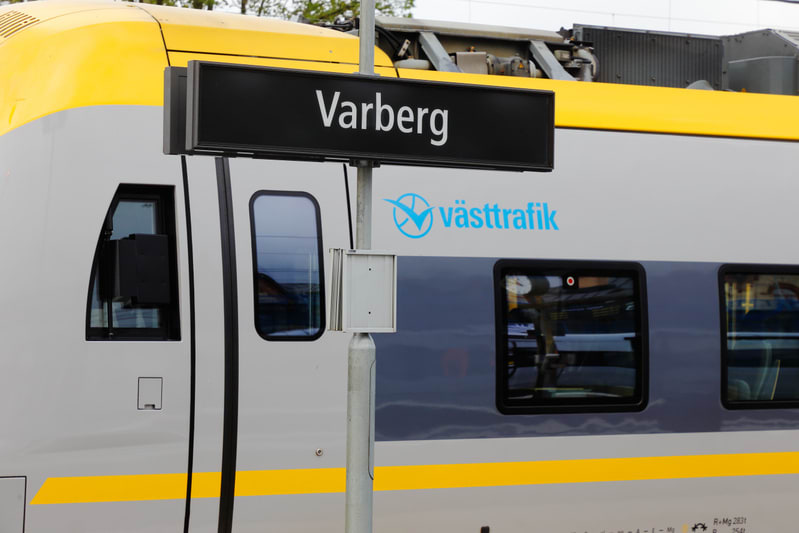 Bilden visar delar av en tågvagn i grått och gult, sannolikt vid en perrong eller station. På vagnens sida syns logotypen för 'Västtrafik' i blått. I förgrunden, mitt i bilden, står en stolpe med en svart skylt som tydligt visar texten 'Varberg' i vi