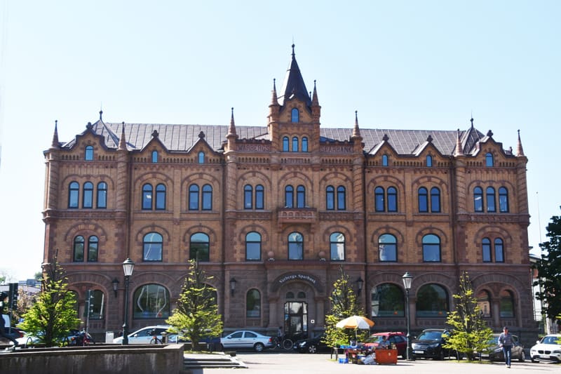 Bilden visar en stor, äldre byggnad i tegel med en karakteristisk arkitektur. Byggnaden har många fönster med rundade överstycken på de nedre våningarna och rektangulära fönster på de övre. Mitt på taket finns ett högt torn med en spetsig spira. Fasa