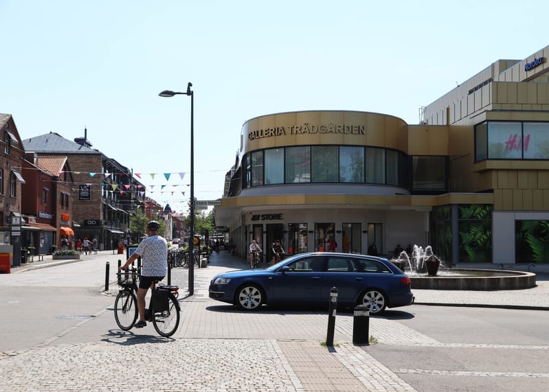 Bilden visar ett gathörn i en stadsmiljö på dagtid. I förgrunden syns en person på cykel i ryggläge. Strax framför cykeln står en mörkblå kombibil parkerad delvis på gatsten. Till höger i bilden dominerar en stor byggnad med guldfärgad fasad och text