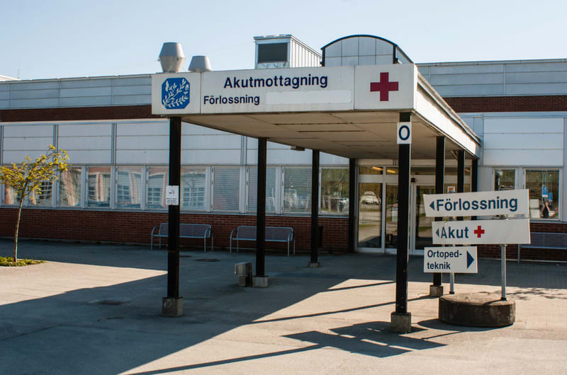 En bild som visar ingången till en Akutmottagning och Förlossning vid ett sjukhus. Ingången är skyddad av ett vitt tak som vilar på svarta stolpar. På taket finns en stor skylt med texten "Akutmottagning" och "Förlossning" samt en röd plus-symbol. På
