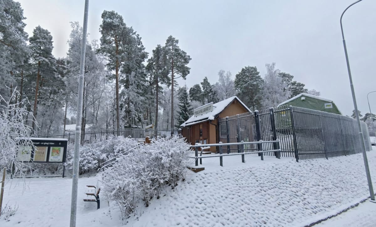 Bilden visar ett snötäckt landskap med barrträd och lövträd, där marken är helt täckt av snö. Ett litet trähus med ett snötäckt tak syns i mitten av bilden, och framför det finns ett svart metallstaket. Till vänster om staketet finns en informationst