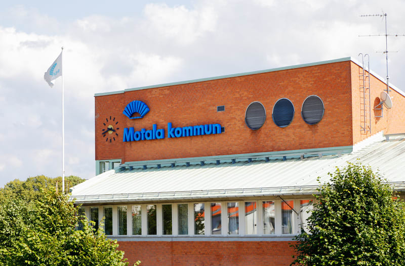En bild som visar en del av en Tegelbyggnad med skylten "Motala kommun" i blå text och logotyp. Ovanför texten syns en blå logotyp formad som ett solfjäderliknande mönster. Till vänster om texten finns en stiliserad urtavla med blå visare. Byggnaden