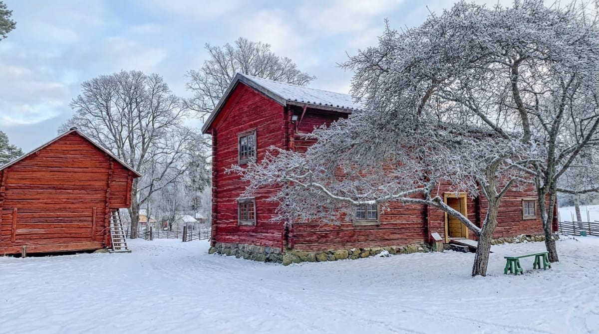 Bilden visar ett vinterlandskap med två rödfärgade trähus täckta av snö. Ett större hus i förgrunden till höger har snö på taket och grenar täckta av rimfrost sträcker sig framför huset. Framför huset står en grön träbänk. Till vänster i bilden syns