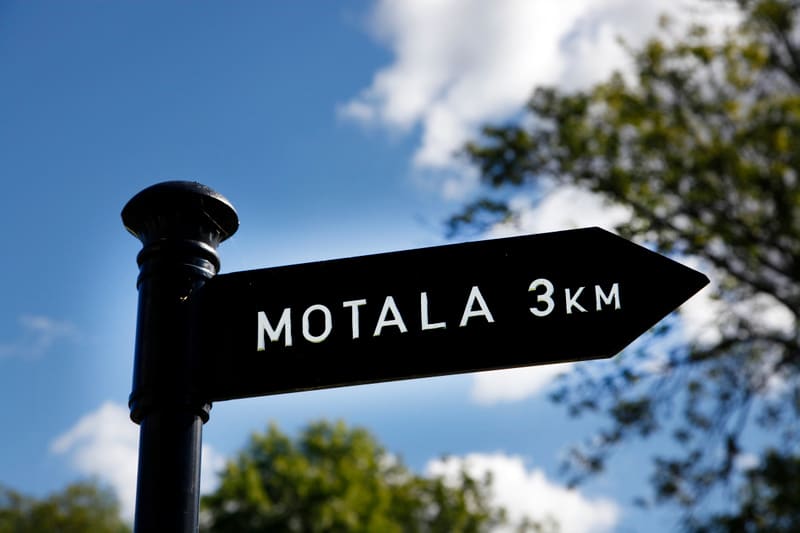 En svart vägvisare av metall med texten "MOTALA 3KM" i vit färg, monterad på en stolpe, syns i förgrunden. Skylten pekar åt höger. Bakgrunden består av en blå himmel med vita moln och oskarpa gröna trädtoppar som indikerar utomhusmiljö och möjligtvis