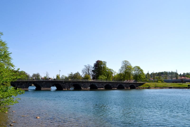 En bild tagen utomhus under dagtid som visar en stenbro med flera valv som sträcker sig över en vattenyta. Vattnet i förgrunden är blåskimrande och reflekterar ljuset. Till vänster i förgrunden syns gröna löv på en gren som hänger in i bilden. Bakom