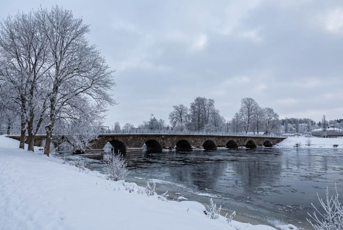En vinterbild visar en bred stenbro med flera valv som sträcker sig över en flod eller älv. Bron har en låg, välvd struktur och synes vara byggd av stenblock. Omgivningen är täckt av snö, och träden längs flodbankarna, samt på avstånd, är snötäckta, vilket ger dem en vitaktig, frostig framtoning. Vattnet i floden är mörkt och reflekterar den gråa himlen, med isflak som flyter på ytan närmare kanterna. På vänstra sidan av bilden syns en snötäckt mark och enstaka buskar. Himlen är mulen och grå, vilket indikerar en kall dag. I bakgrunden syns skogklädda kullar eller höjder även dessa snötäckta, samt en antydan till bebyggelse eller en annan bro längre bort till höger.