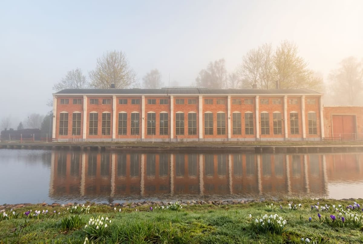 En stor, röd tegelbyggnad med många fönster sträcker sig horisontellt längs en vattenyta. Byggnaden reflekteras tydligt i vattnet framför den. Ovanför byggnadens tak kan man se kala trädgrenar och disig luft. I förgrunden, på marken närmast betraktar