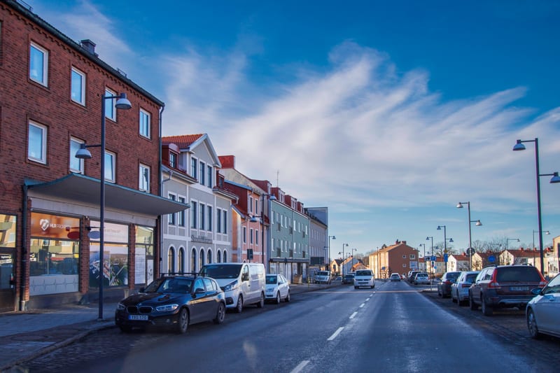 Bilden visar en gata i en stadsmiljö under dagtid. På vänster sida av gatan syns en rad flervåningshus i olika färger, bland annat rött tegel och ljusare putsfasader. På markplan i ett av husen finns en butik med stora skyltfönster. Framför dessa hus