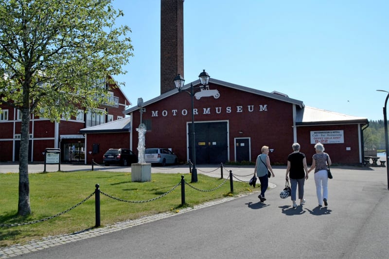 En bild tagen utomhus under dagtid, som visar en röd byggnad med texten "MOTORMUSEUM" på fasaden. På taket av byggnaden syns silhuetten av en motorcykel. Till vänster om museibyggnaden finns en skorsten av tegel och en annan röd byggnad med balkonger