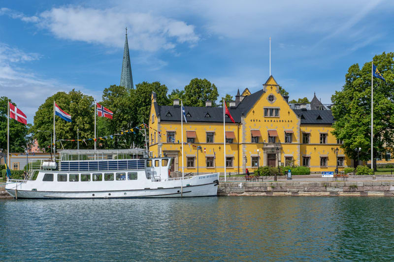 Bilden visar ett landskap med en massa vatten i förgrunden och byggnader och träd i bakgrunden. I vattnet ligger en vit passagerarbåt förtöjd. På båten syns flaggor från Danmark, Nederländerna och Norge. I bakgrunden till höger står en stor, gul bygg