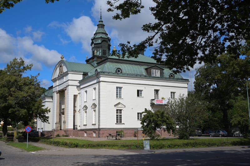 Bilden visar en stor, ljus byggnad med en grön kupol och ett torn med en spira, under en blå himmel med vita moln. Byggnaden har vita väggar, flera fönster i olika storlekar och ett grågrönt tegeltak. En del av byggnaden har kolumner vid entrén och e