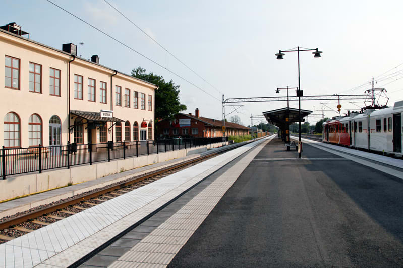 Bilden visar en järnvägsstation från en låg vinkel. Till vänster syns en ljusgul byggnad med bruna fönsterramar och ett svart tak. Framför byggnaden finns en svart metallstaket och en perrong vid järnvägsspåret. Ett skylt med texten "MOTALA" är synli