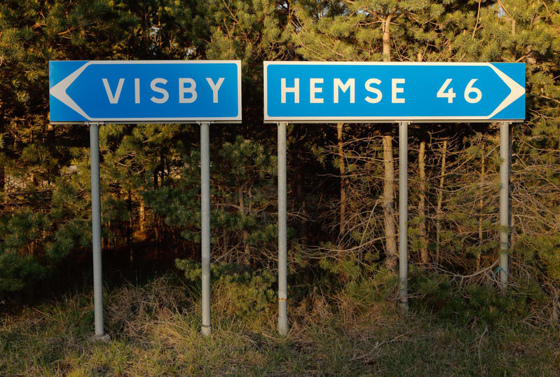 Bilden visar två blå vägskyltar med vit text och pilar, placerade bredvid varandra. Skylten till vänster pekar åt vänster och har texten "VISBY". Skylten till höger pekar åt höger och har texten "HEMSE 46". Båda skyltarna är monterade på grå metallst