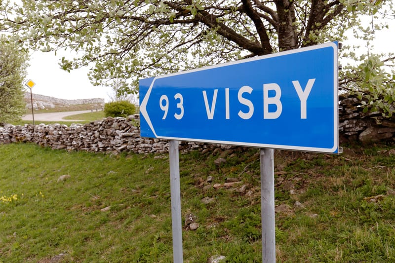 Bilden visar en blå vägskylt med vit text som pekar åt vänster. På skylten står det "93 VISBY". Skylten sitter monterad på två grå stolpar. I bakgrunden syns en stenkant längs en gräsklädd sluttning. Till vänster om skylten syns en kurvig grusväg och