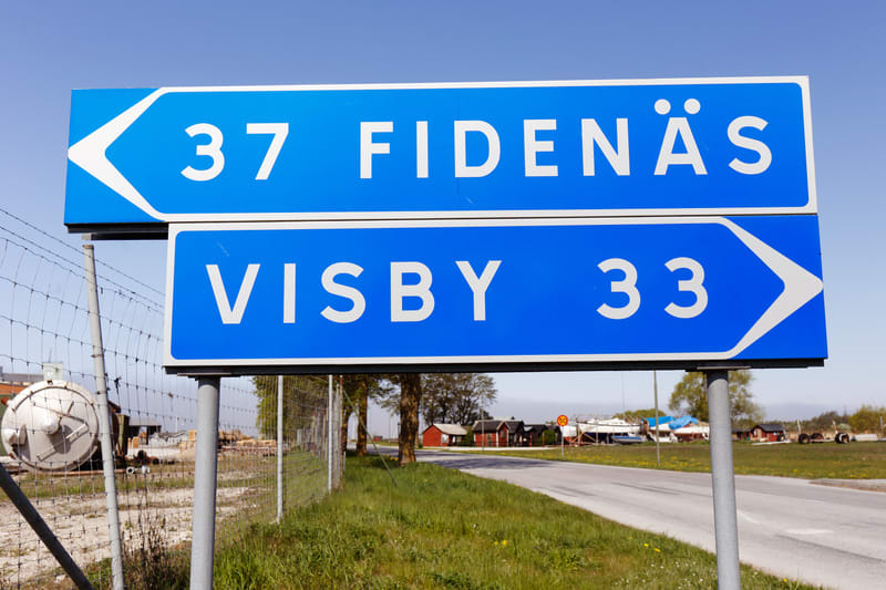 En bild tagen utomhus under dagtid som visar en blå vägskylt monterad på två stolpar. Den övre delen av skylten har texten "37 FIDENÄS" med en vit pil som pekar åt vänster. Den nedre delen av skylten visar "VISBY 33" med en vit pil som pekar åt höger