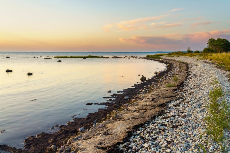 Bilden visar en havsvik med en strandlinje. Vattnet, som reflekterar himlens färger, är lugnt och färgerna går från ljusblått vid horisonten till en gulaktig ton närmare stranden. Ett flertal mörka stenar syns utspridda i vattnet. Stranden består av