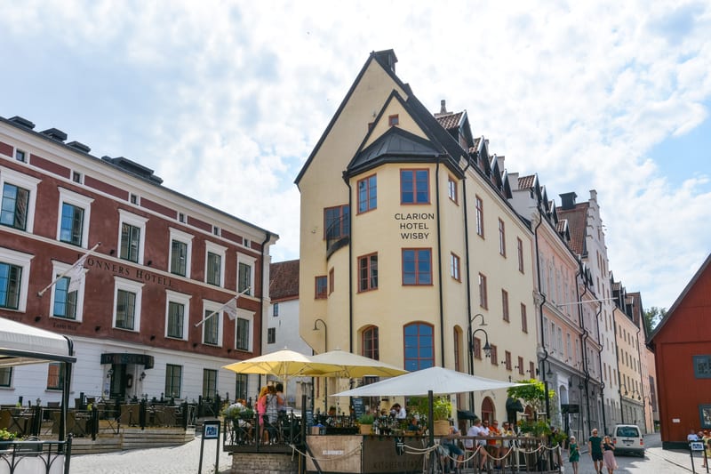 Bilden visar en gatuscen med flera byggnader i varierande färger, en av dem identifieras som "Clarion Hotel Wisby" och en annan som "Donners Hotell". Det är en solig dag med delvis molnig himmel. Framför hotellen finns uteserveringar med människor so