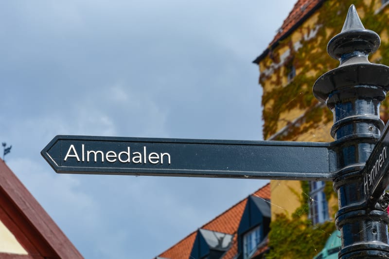 En svart metallskylt med texten ”Almedalen” i vit färg pekar mot vänster. Skylten är fäst vid en svart, dekorerad stolpe där en annan skylt med texten ”Hamnen” delvis är synlig längre ned till höger. I bakgrunden syns delar av äldre byggnader med fas