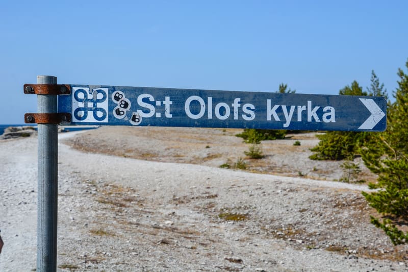 Bilden visar en blågrå skylt fäst vid en metallstolpe. Skylten har vit text "S:t Olofs kyrka" med en vit pil som pekar åt höger. Det finns även klistermärken synliga på skylten, varav ett är en vit symbol på en svart bakgrund som liknar en fyrkantsym